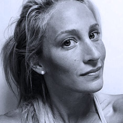 A woman with long blonde hair, light skin, and wearing a small stud earring looks at the camera with a slight smile against a plain background. The image is in black and white.